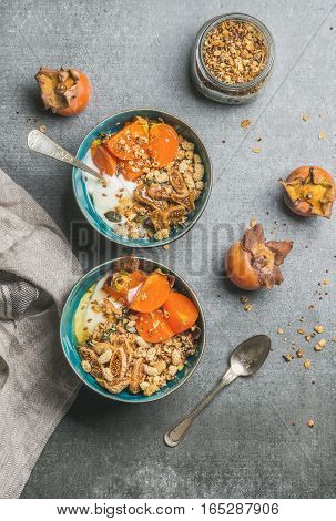 Healthy vegetarian breakfast. Oatmeal, quinoa granola with yogurt, dried fruit, seeds, honey, fresh persimmon in blue ceramic bowls over grey background, top view. Dieting, allergy-friendly concept
