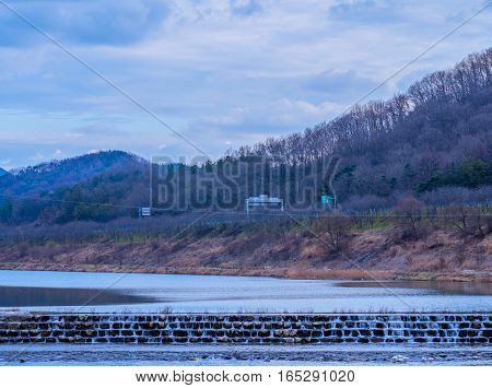 Rural landscape of small waterfall in a river with foothills and a country road in the background and a beautiful blue sky with puffy white clouds