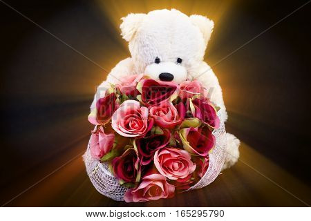 Selective focus at bouquet of beautiful artificial red roses in the arms of blurry white bear doll on black background and ray effect concept love for valentine day or spacial anniversary