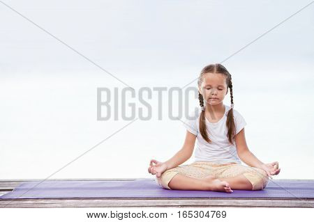 Child doing meditating exercise on wooden platform sea shore outdoors. Healthy lifestyle. Yoga girl