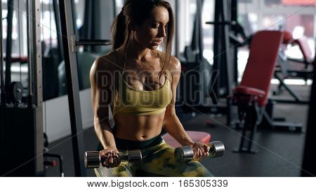 Brunette lifting dumbbell and working on her biceps in a gym. Young lady with beautiful body. Determined sexy woman working on own shape.