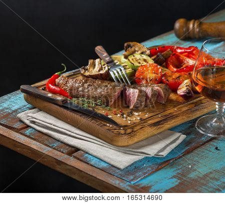 Steak grilled and grilled vegetables on a wooden board with a glass of brandy on a dark background