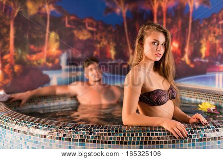 Young beautiful romantic couple relaxing together in jakuzzi at spa centre, woman posing to camera