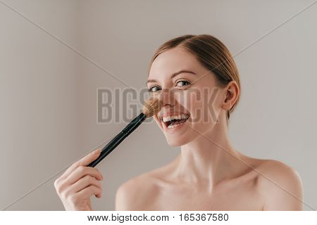 Going crazy. Attractive young woman looking at camera and touching her nose with make-up brush while standing against background