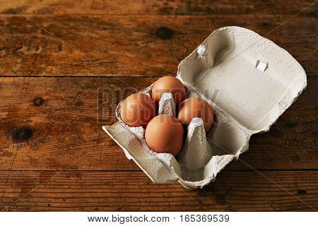 Open egg carton for six eggs containing four brown eggs on a rough rustic brown wooden table