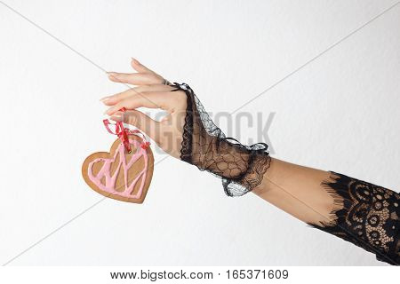 One beautiful female hand in black lace glove holds a heart shaped gingerbread cookie on red ribbon against white beackground horizontal view. Valentine's day and love concept