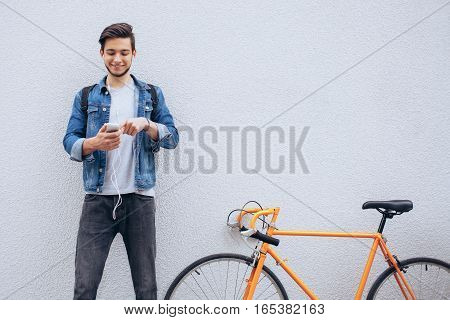 Young student in blue jeans jacket standing near the orange bicycle looking at phone and listening to music. He is scrolling list of his favourite tracks. Wall background