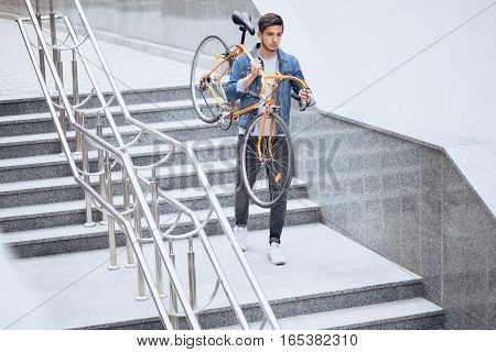 The guy in the blue jeans jacket carrying on his shoulder orange bike. A young handsome man with beard holding it on his right arm. He is going down stairs at work. Urban background