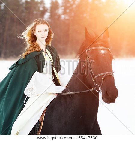 Young girl in white dress and green cape riding horse on snowy field near forest