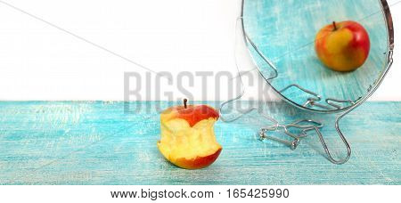Bit of apple and reflection in a mirror of the whole apple. The taken a bite apple is reflected in a blue colored table as whole. At the left above empty seat white