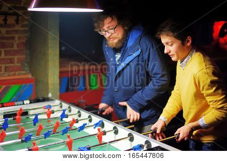 Two young men in evening bar play kicker. A friend with a beard and the other in the yellow jacket. They are concentrated.