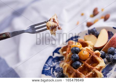 Closeup of a fork cutting through a blueberry waffle decorated with fresh blueberry in a nice dish.