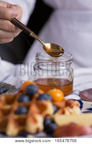 Blueberry apple waffle with honey set on a dish with a chef's hand holding spoon for a jar of honey ready to serve.