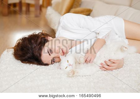 Cute curly girl in a white silk dressing gown in the early morning playing with white fluffy cat sitting on sofa