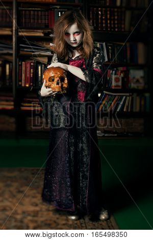 Little girl vampire in library holding in hand the human skull