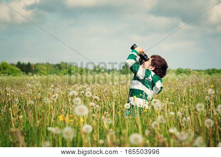 Child wathing in benoculars on the field with dandelions. Boy playing on the meadow in summer