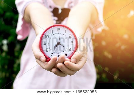 Holding red clock on hand and giving this to you with garden background.