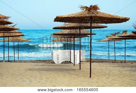 Empty beach in the seaside resort with folded sun loungers and wooden sheds with thatched roof on a background of blue sea.