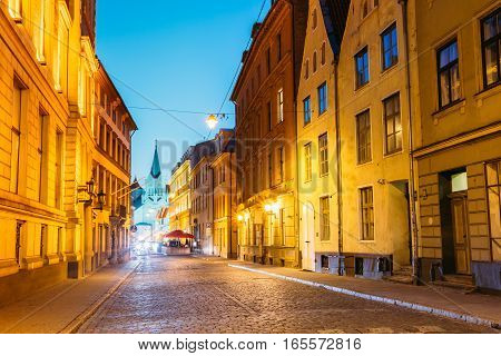 Riga Latvia. The White Tower With Pyramidal Spire Of Our Lady Of Sorrows Or Virgin Of Anguish Church, Ancient Catholic Church On Pils Street In Evening Illumination Under Summer Blue Sky