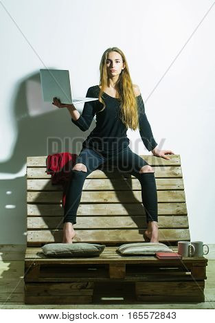 Pretty girl or beautiful woman cute sexy fashion model with long hair in casual clothes with laptop computer sits on backrest of wooden pallet sofa on white wall