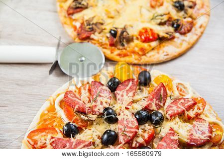 Pizza cutter together with two large pizzas with cheese olives and salami mushrooms and tomatoes. On a wooden stand.
