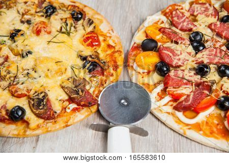 Pizza cutter together with two large pizzas with cheese olives and salami mushrooms and tomatoes. On a wooden stand.