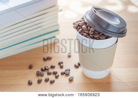 A paper cup of full roasted aroma coffee beans with blurred background of some fallen coffee beans on wooden board business concept.