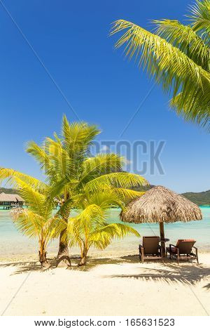 Two sunlounger chairs under a thatched parasol on a sand beach with a view on the lagoon and Mount Otemanu on the tropical island of Bora Bora near Tahiti in French Polynesia. Vertical composition