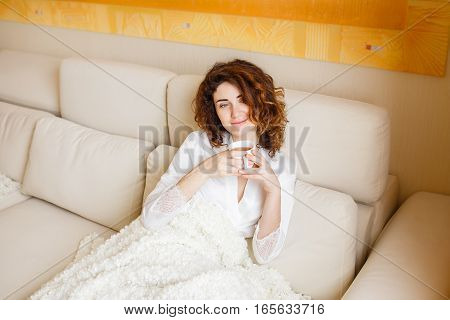 Pretty curly hair girl in a white silk dressing gown in the early morning drinking coffee