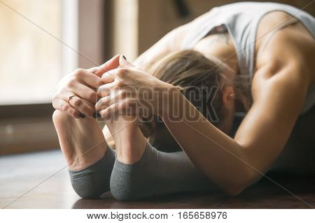 Close up of young woman practicing yoga, sitting in Seated forward bend exercise, paschimottanasana pose, working out, wearing sportswear, grey pants, bra, indoor, home interior background, horizontal