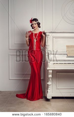 Young Beautiful Girl In Red Dress Style Of The 20's Or 30's With Glass Of Martini Near The Piano. Vi