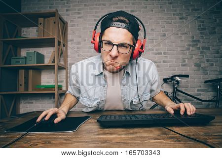 Young man with eyeglasses and earphones playing game at home and streaming playthrough or walkthrough video. He is really concentrated on gaming. His face expression is aggressive