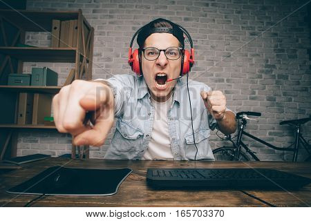 Young man in red headphones playing game at home and streaming playthrough or walkthrough video. Crazy guy is shouting because of unjustice in virtual game and pointing at the screen with one finger