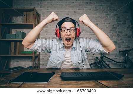 Young man playing game at home and streaming playthrough or walkthrough video. Attractive man is wearing eyeglasses and earphones. He is happy to win this fight in vr. Boy lifting his fists above the head