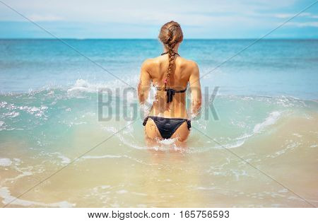 Outdoor summer portrait of young elegant fit woman standing backwards and going into sea to swim in waves near tropical beach. Sports girl in bikini enjoy nature paradise summer vacation in Thailand