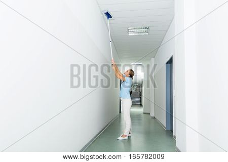 Young Woman Cleaning The Ceiling With Mop On The Corridor Of The Building