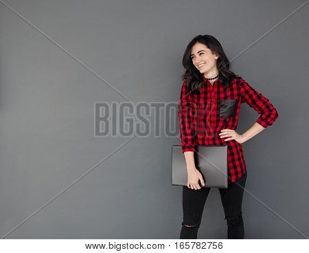Full length portrait of a smiling young woman standing and holding laptop computer while looking at copy space isolated on a grey background
