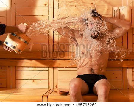 Excited man or bather with muscular sexy torso body wet with big splash of water from bucket in sauna thermal bath on wooden background