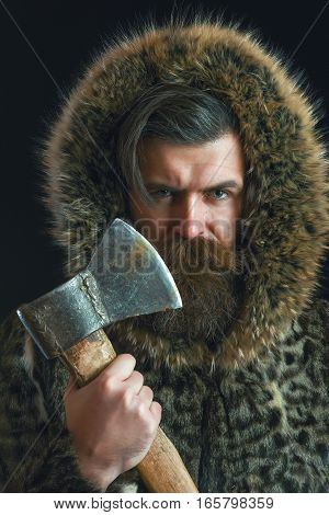 handsome bearded man or woodman guy with fashionable mustache and beard on serious face in brown fur coat holds sharp axe or ax on black background