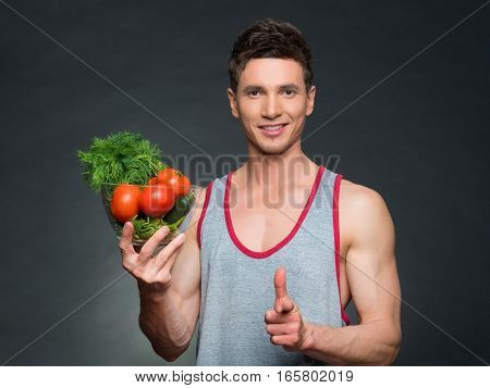 Young fittness trainer and nutritionist holding a bowl of vegetables, on black background