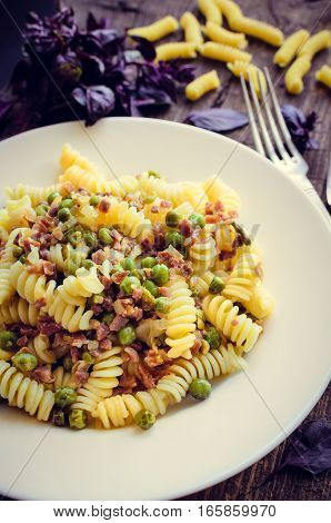 Fusilli with prosciutto and green pease on old wooden background. Traditional Italian pasta with fresh basil. Selective focus.