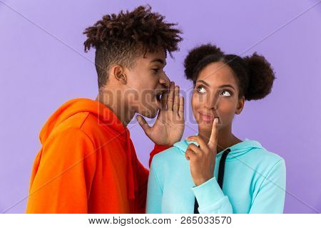 Photo of beautiful african american man talking in ear of young woman isolated over violet background