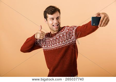 Portrait a smiling young man standing isolated over beige background, taking selfie with mobile phonem, showing thumbs up