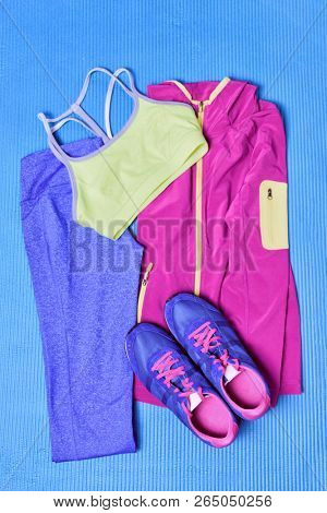 Pilates or yoga activewear fashion outfit to go train in gym . Fitness clothes on exercise mat with running shoes and blue leggings, cute girl clothes on background, top view.