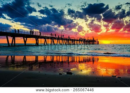 People Stroll On Glenelg Jetty At Sunset Sky Splashed With Colourful Hues Of Red, Orange And Blue In