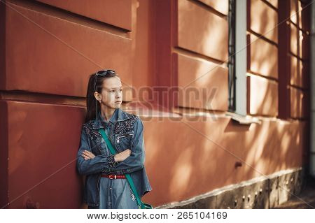 Serious Little Girl In Jeans Clothes Stands Near An Old Restored Beautiful Building.