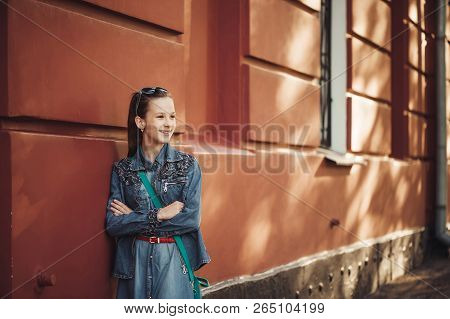 Cheerful Little Girl In Jeans Clothes Stands Near An Old Restored Beautiful Building.
