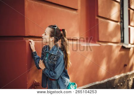 Curious Little Girl In Jeans Clothes Stands Near An Old Restored Beautiful Building.