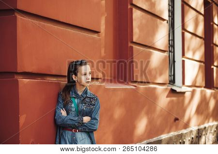 Serious Little Girl In Jeans Clothes Stands Near An Old Restored Beautiful Building.