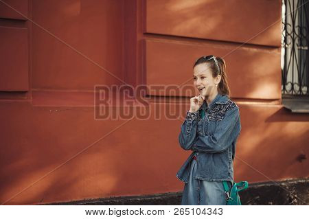 Cheerful Little Girl In Jeans Clothes Stands Near An Old Restored Beautiful Building.